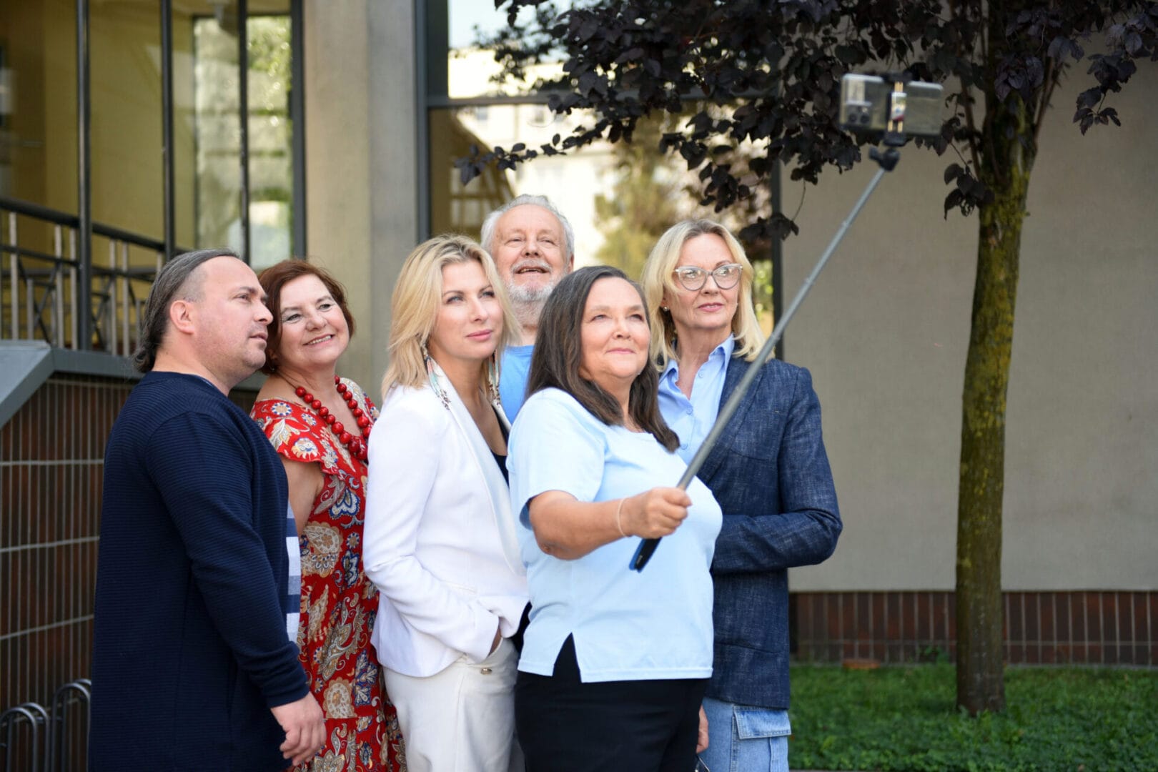 Grupa sześciu os&oacute;b robi selfie na zewnątrz. Kobieta na pierwszym planie trzyma kijek do selfie, reszta pozuje uśmiechnięta.