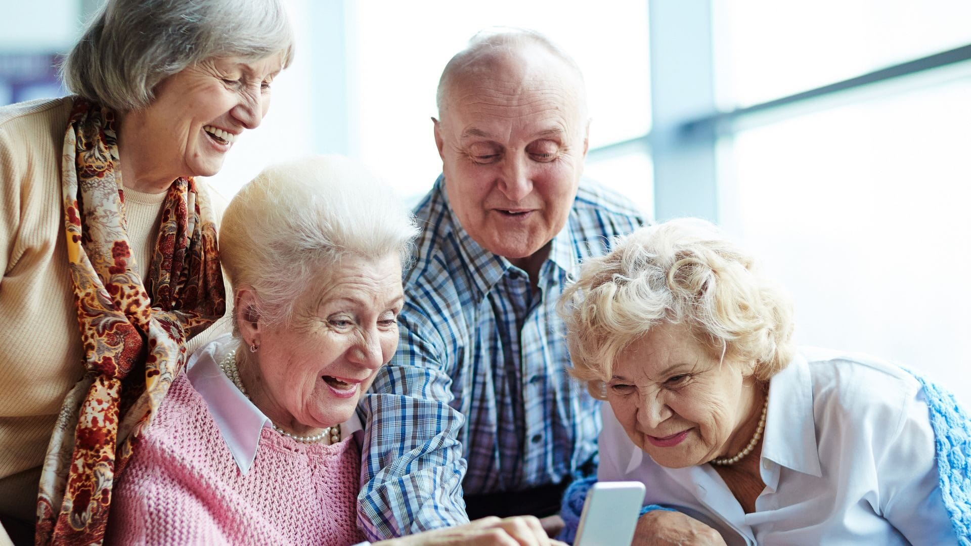 Grupa czterech uśmiechniętych seniorów - trzy kobiety i jeden mężczyzna - pochylających się nad smartfonem, któremu przyglądają się z zainteresowaniem.