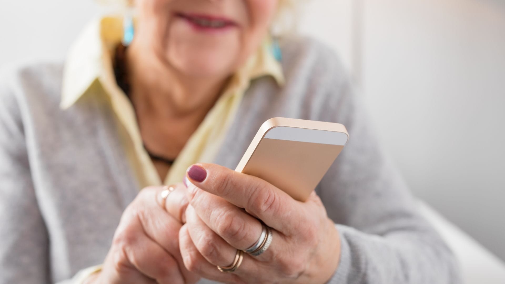 Zbliżenie na dłonie starszej kobiety trzymającej smartfona. Widoczne są pierścionki na palcach i manicure na paznokciach.