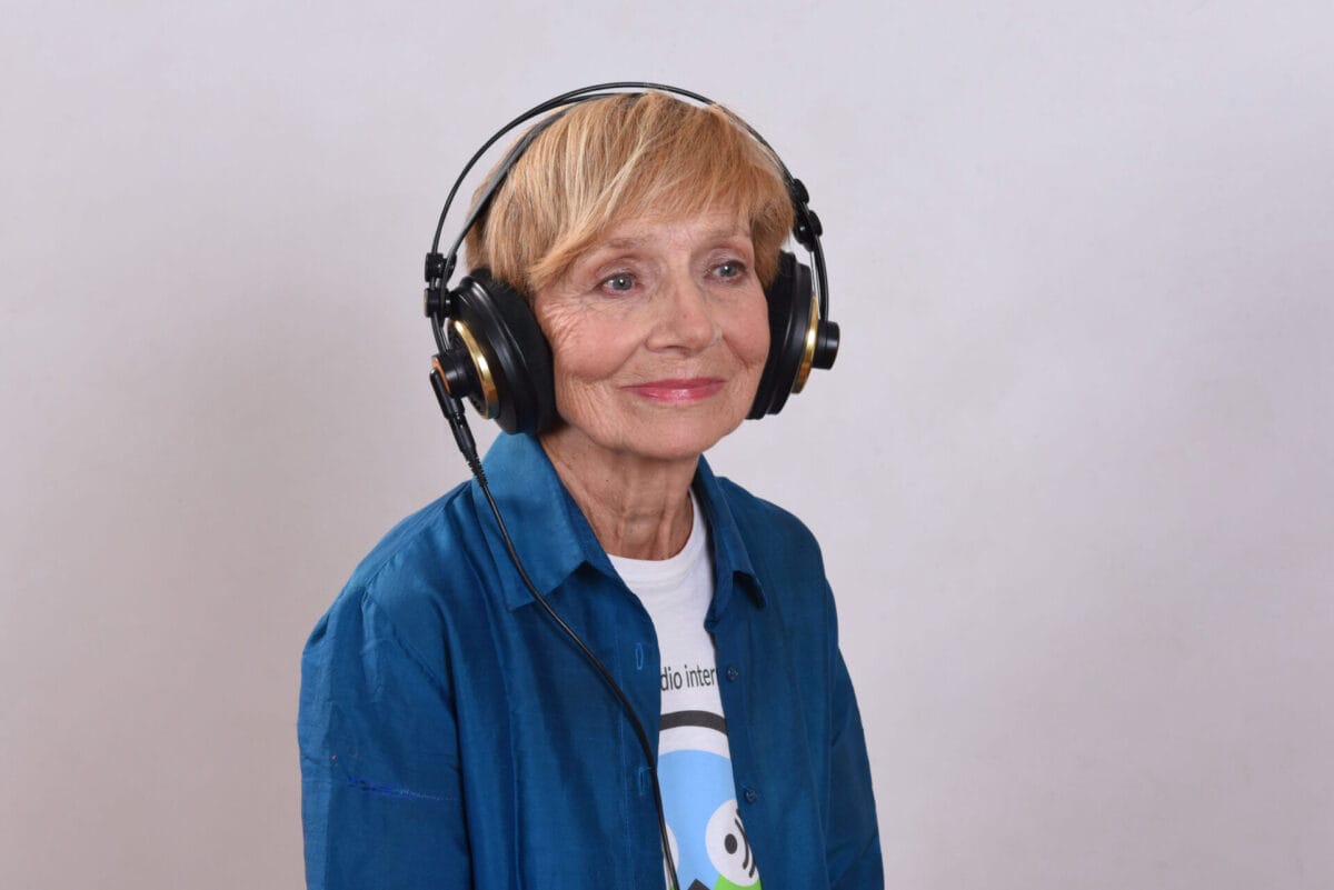 Seniorka w słuchawkach studyjnych, uśmiechnięta, na jasnym tle.