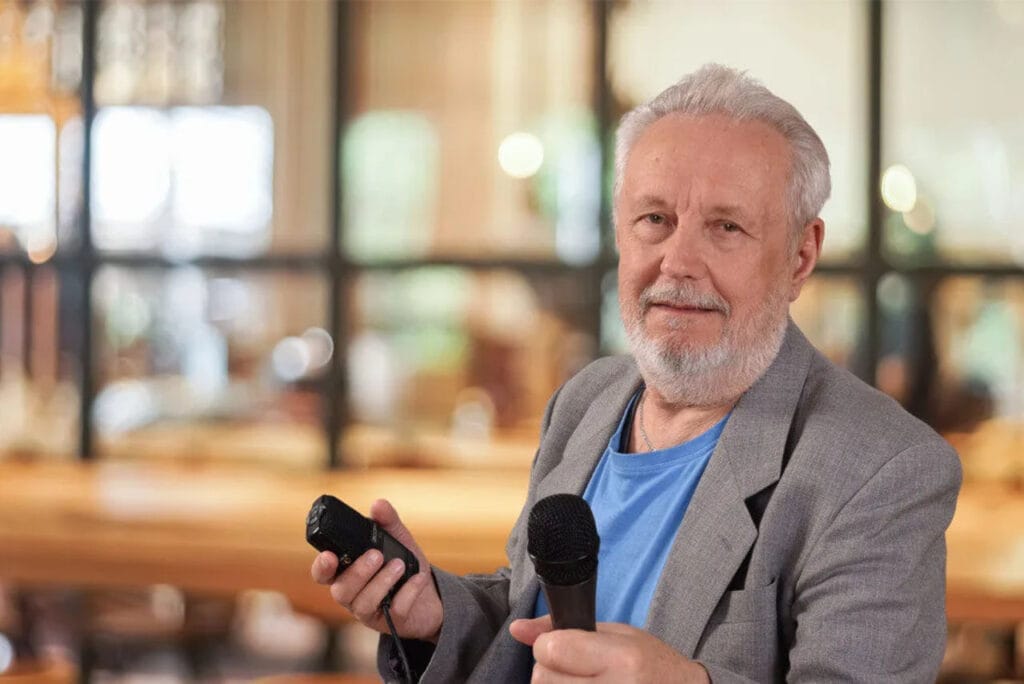 "Starszy mężczyzna z siwą brodą i włosami, ubrany w szary marynarkę i niebieską koszulkę, trzymający mikrofon i małe urządzenie nagrywające. Stoi w pomieszczeniu z rozmazanym, jasnym tłem